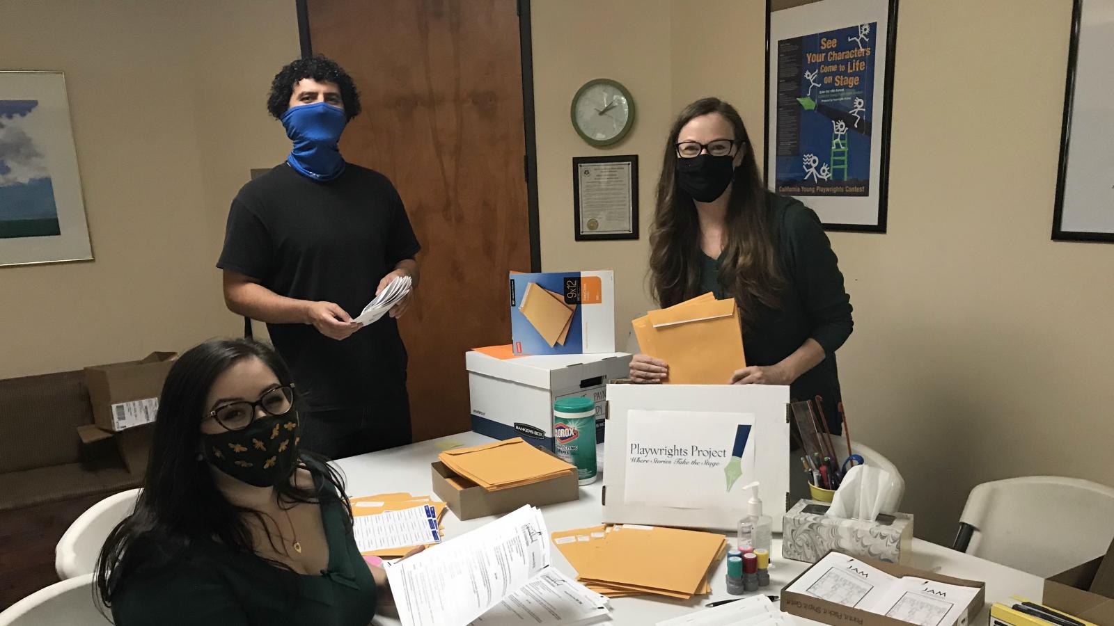Three people wearing masks look at the camera as they prepare large envelopes for mailing