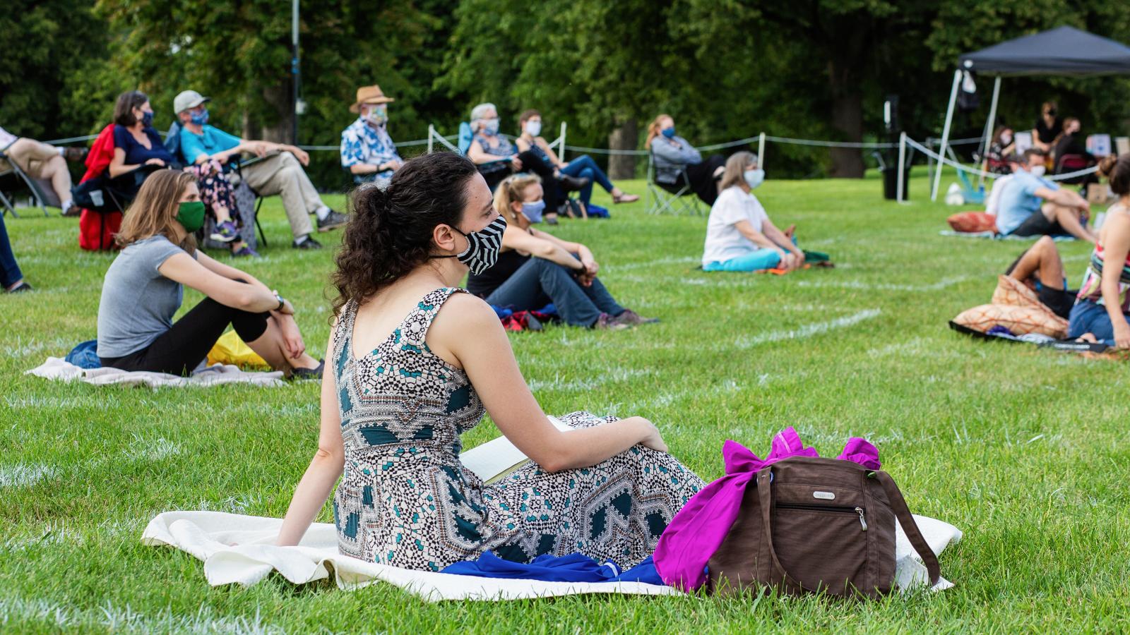 People wearing masks sit on picnic blankets on the grass while spread out from one another