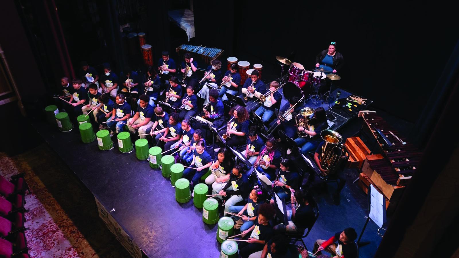 Overhead shot of children on stage with musical instruments, the front row students banging on plastic tubs. 