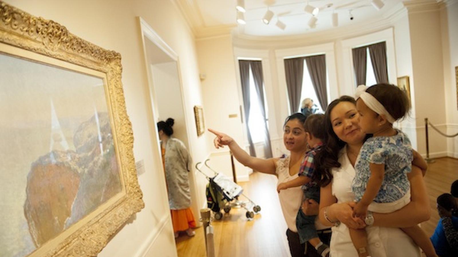 two women holding their toddlers and looking at a landscape painting on the wall of a museum