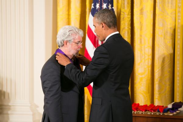 Man with gray hair and gray beard receiving a medal from tall black man in a suite in front of an American flag. 