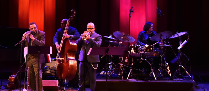 A flutist, upright bassist, trumpeter, and drummer all perform onstage.