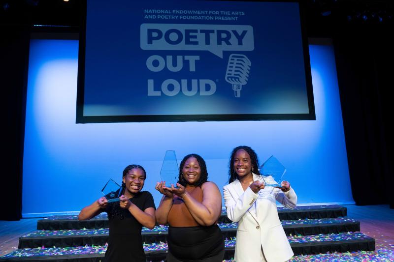 Three young Black women stand in front of Poetry Out Loud sign holding trophies aloft