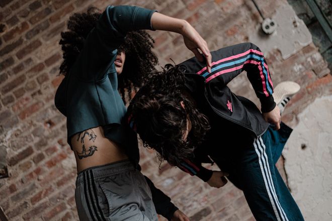 Two dancers improvise during a rehearsal. One stands at an angle with her arm curved above her while another dancer immediately in front of her stands in profile, curling her body forward.