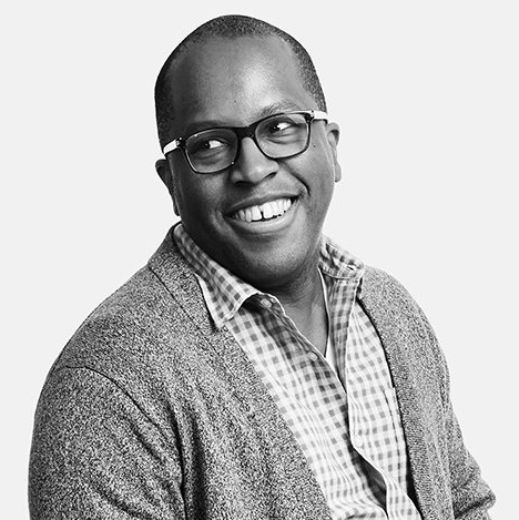 Headshot of a Black man smiling and wearing glasses, a checkered button-down shirt, with a jacket over the button-down shirt.