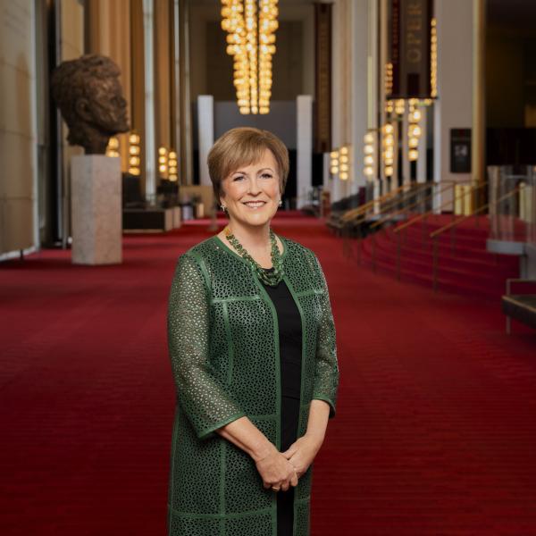 photo of Deborah F. Rutter, a middle-aged white woman with short brown hair. She wears a green loosely woven long jacket over a black dress and stands in the main hall of the Kennedy Center