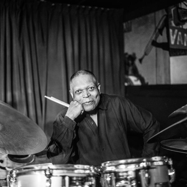 Black and white photo of a man sitting behind a drum kit holding sticks in his right hand. 