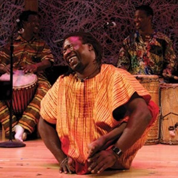 Man in bright African garb dancing without using legs onstage in front of drummers