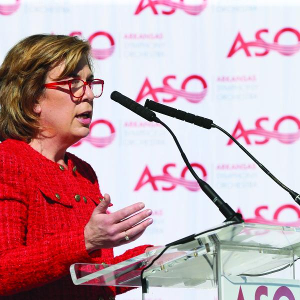 A woman speaks at a podium, a step-and-repeat banner is behind her which shows the logo for the Arkansas Symphony Orchestra.