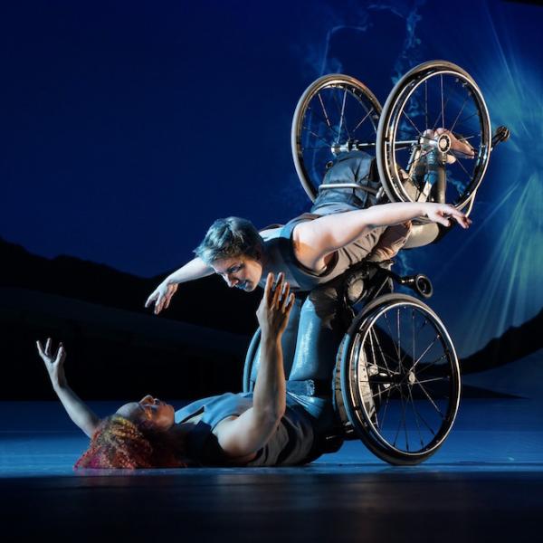 Laurel Lawson, a white woman with short cropped teal hair, is flying in the air with arms spread wide, wheels spinning, and supported by Alice Sheppard. Alice, a multiracial Black woman with coffee-colored hair, is lifting from the ground below. They are making eye contact and smiling. A burst of white light appears in a dark blue sky. 