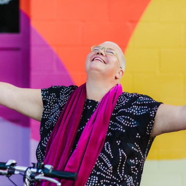 Petra Kuppers, a white queer disabled cis woman of size with yellow glasses, shaved head, pink lipstick and a black dotted top, smiles up to the sky, arms outstretched, embracing the world. Her mobility scooter’s handlebar is visible at the bottom of the image. She is in front of a multicolored wall: purple, pink, yellow and orange.