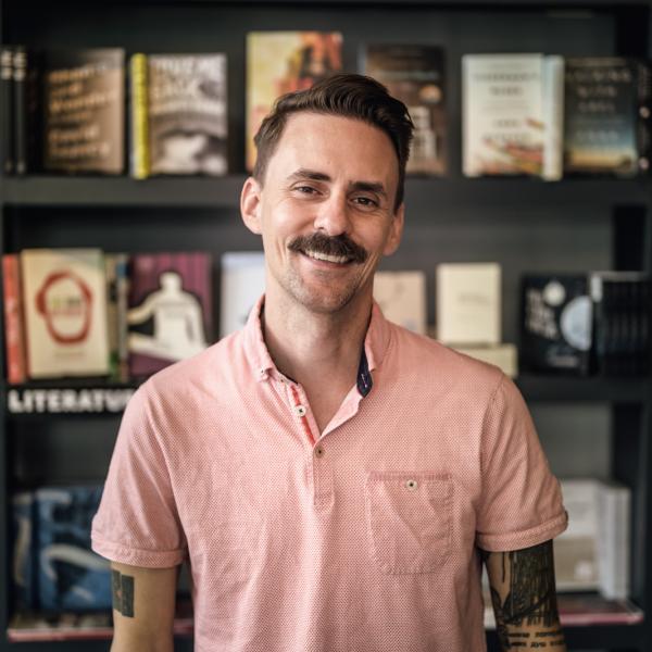 A white man with short brown hair and a moustacha wearing a short sleeved light red collared shirt smiles in front of a bookshelf