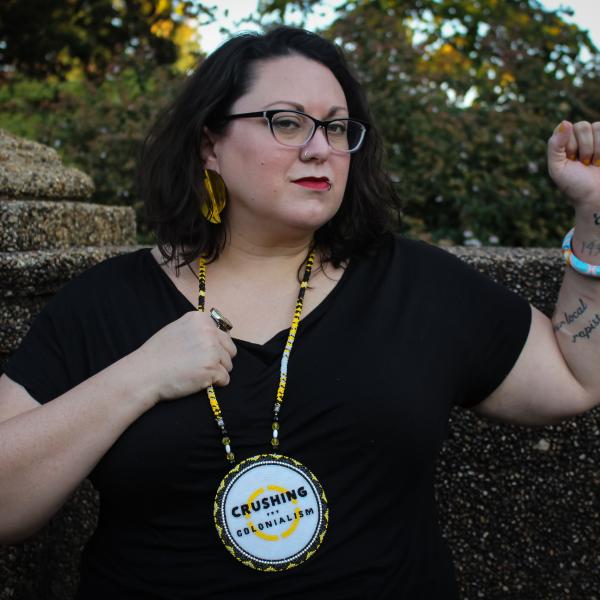 a Native woman with short wavy hair wearing glasses and a large beaded pendant that says Crushing Colonialism