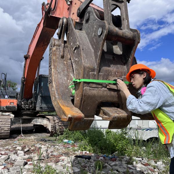 A person wearing an orange hard hat and a yellow safety vest attaches a camera to the claw of a large construction excavator. The claw has a green ribbon around it, and the person is leaning forward, peering through the camera's viewfinder.