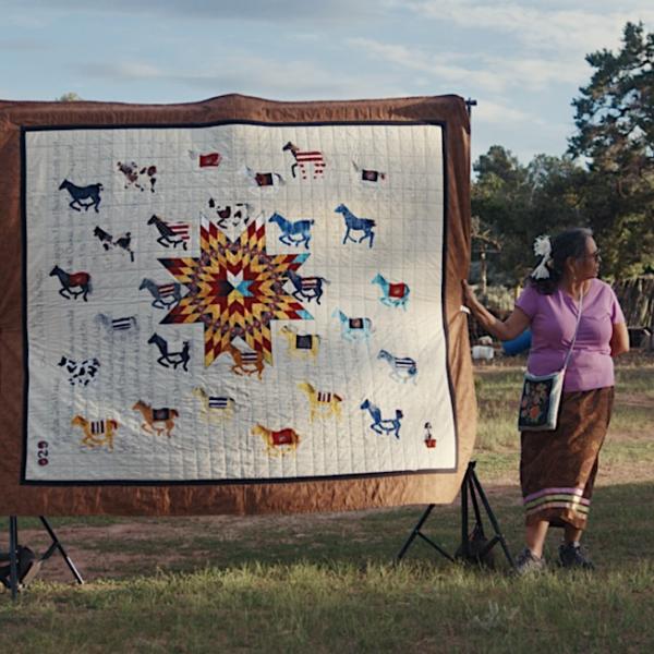 Susan Hudson standing outdoors with a large quilt displayed on a frame