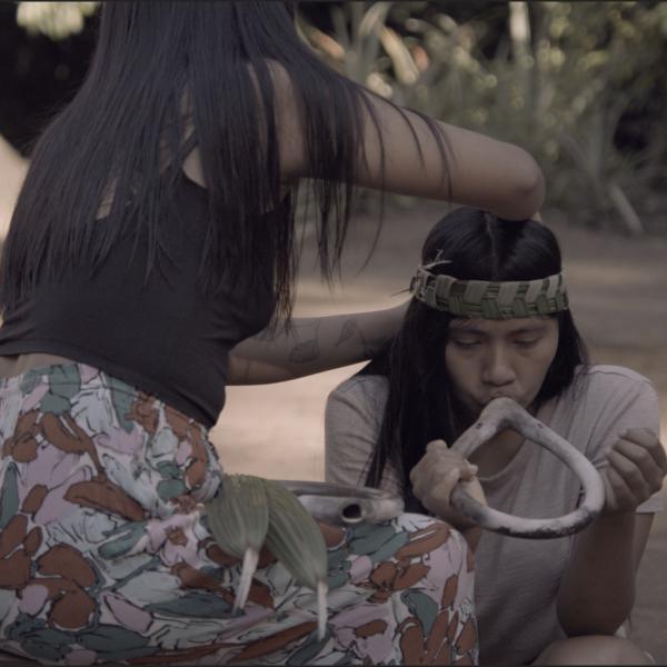 Film still of two women: Woman on left is tying a headpiece on to the woman on the right. Woman on the right is kneeling and playing an instrument.