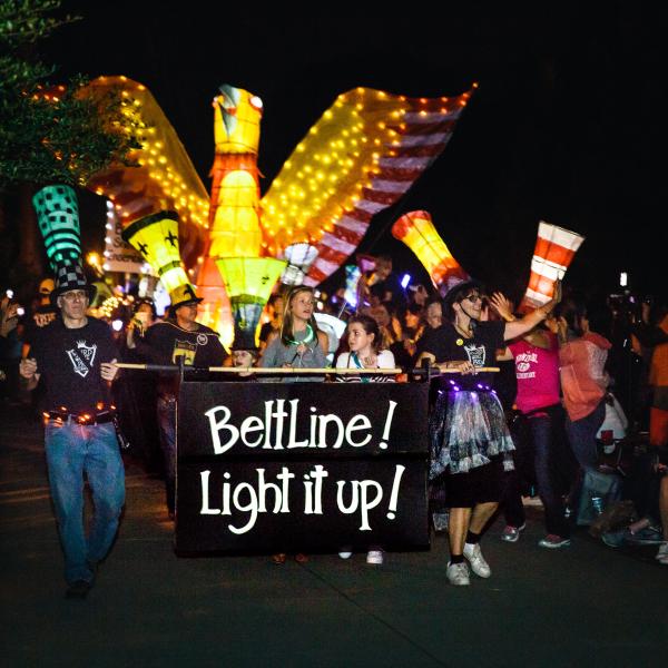 People lead a parade at night, holding a large sign that says "Beltline! Light it up!" They, as well as several others, are wearing tall hats that look like illuminated paper lanterns. Behind them is a large illuminated paper lantern of an orange and yellow bird.