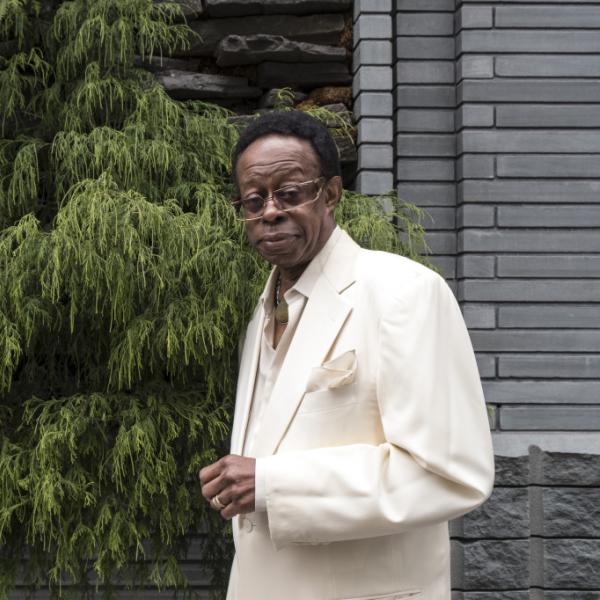 Older Black man in a white suit outside in front of a house with a tree nearby.