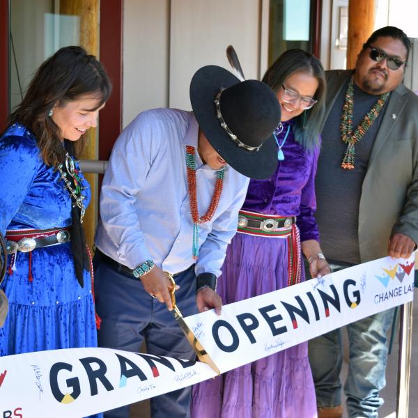 A small group of people stand behind a banner reading "Grand Opening". A man in the center is cutting the banner with large golden scissors.