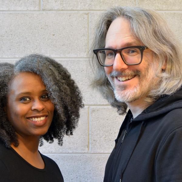 photo of a Black woman with long curly hair and a white man with longish hair, beard and glasses with a cinderblock wall in the background