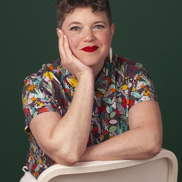 Headshot of a White woman wearing a multi-colored floral shirt with a solid green background behind her
