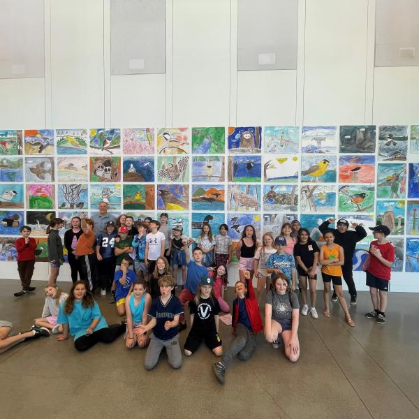 A group of students and one male adult stand in front of a gallery of artwork.