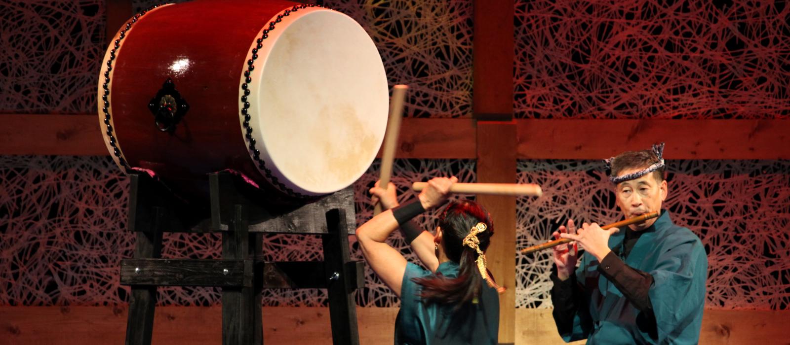 Woman banging a large drum while man plays flute on stage. 