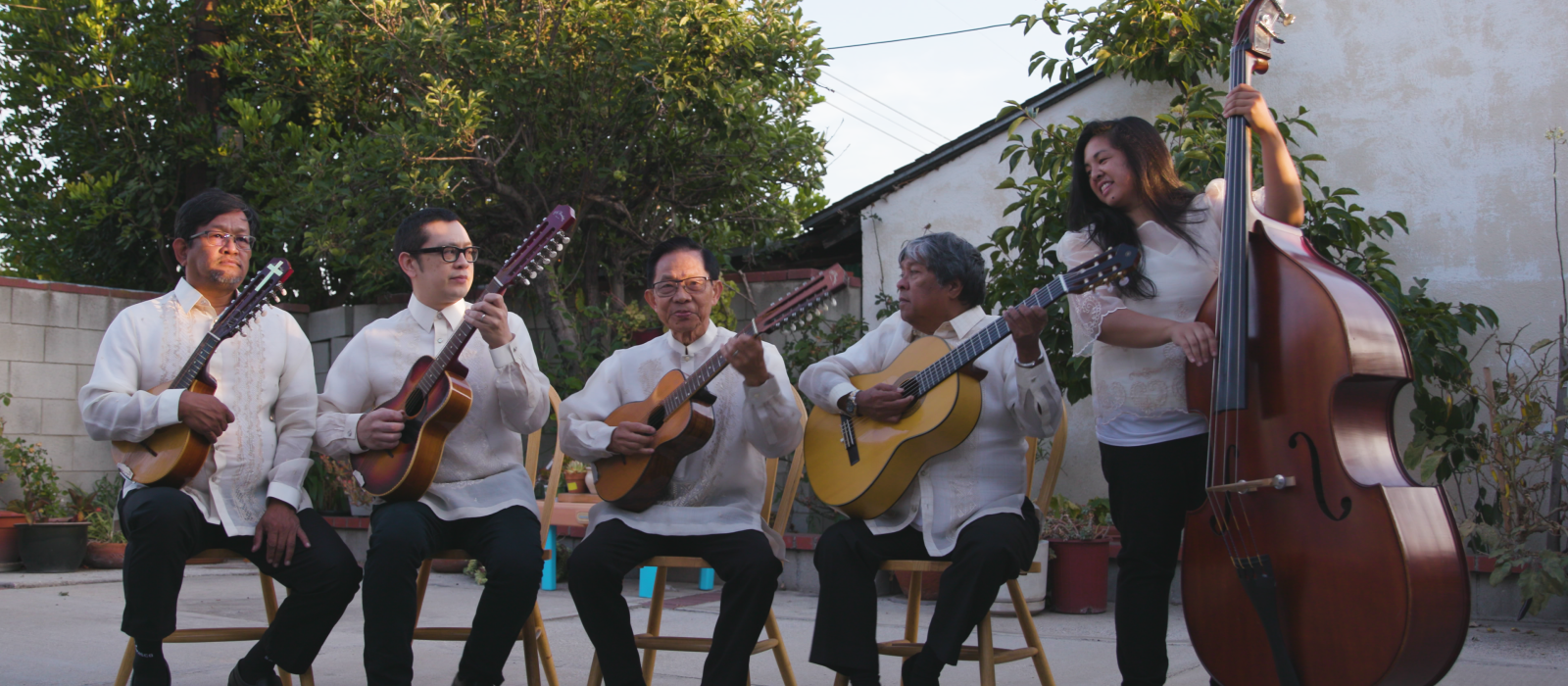 Group of musicians holding instruments seated outside all wearing white shirts. 