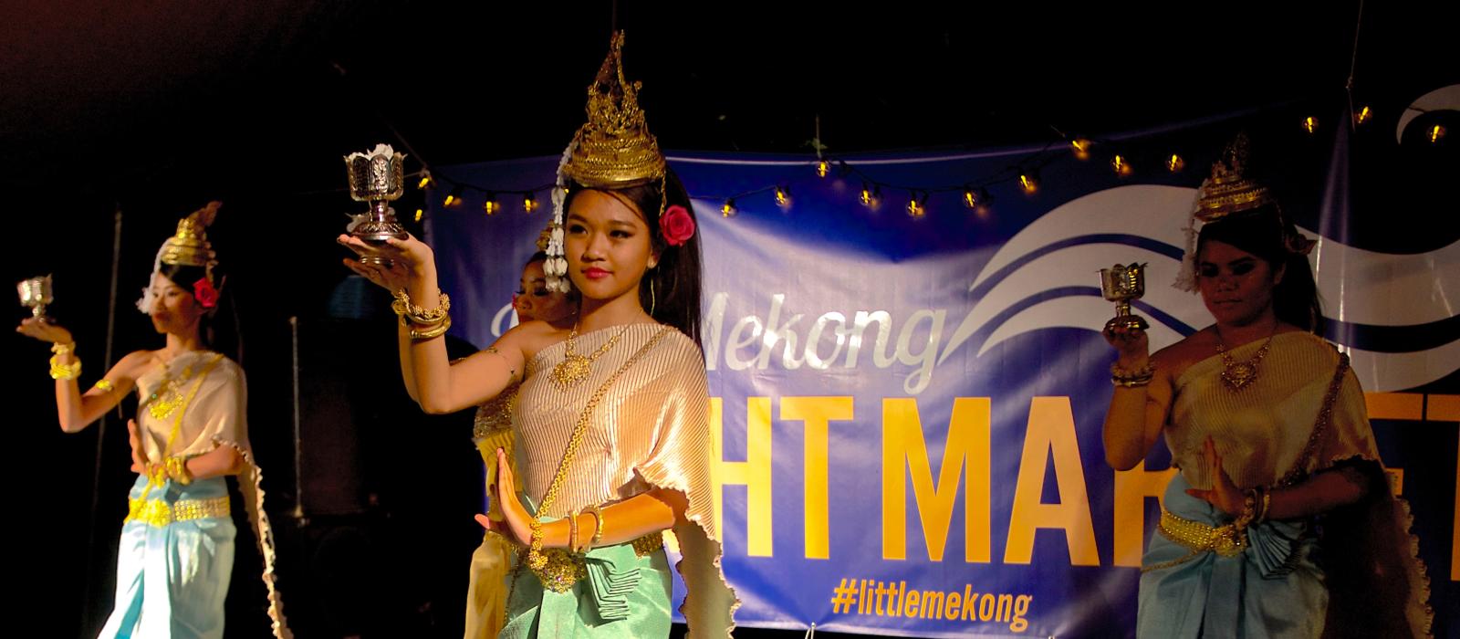 Women in Cambodian garb of blue and yellow in parade down the street, in front of a banner. 