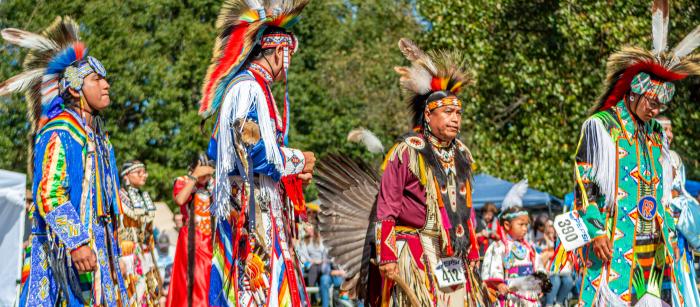 A group of four men in traditional regalia walking in a line outdoors.
