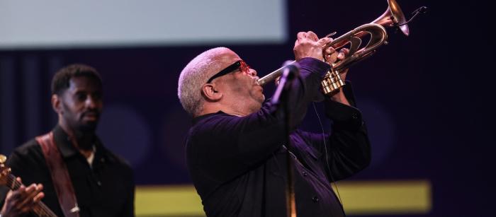 A man with glasses performs on the trombone. In the background behind him is a musician on the bass guitar.