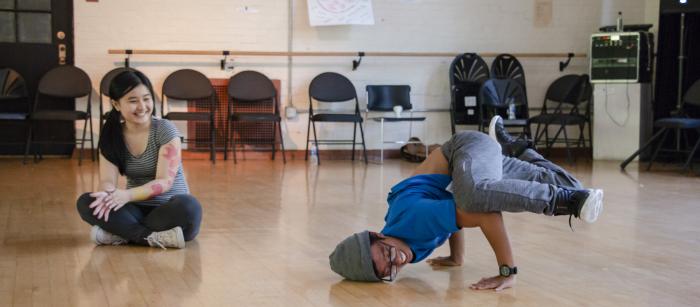 Student wearing hat dancing while a girl sitting cross-legged nearby watches. 