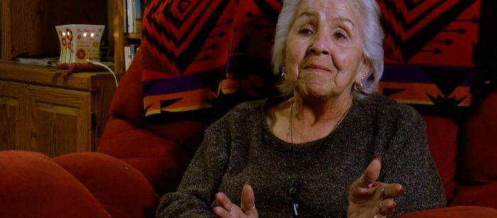 Woman with gray hair wearing dark sweater sitting on red, pillowed chair with Native-designed blanket behind her head.