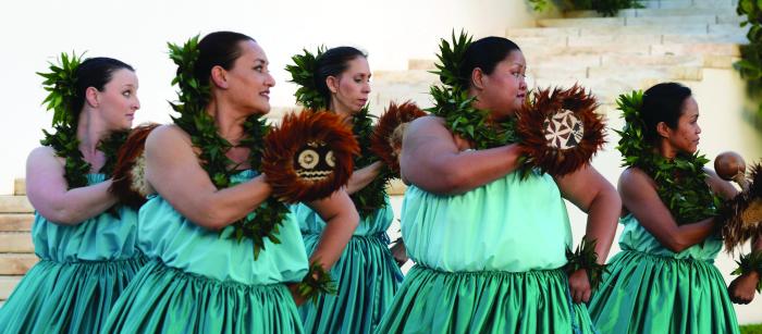 Women in blue dresses holding feathered gourds perform a traditional Hawaiian dance.