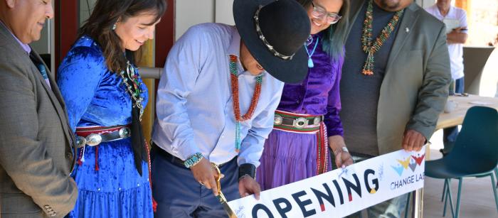 A small group of people stand behind a banner reading "Grand Opening". A man in the center is cutting the banner with large golden scissors.