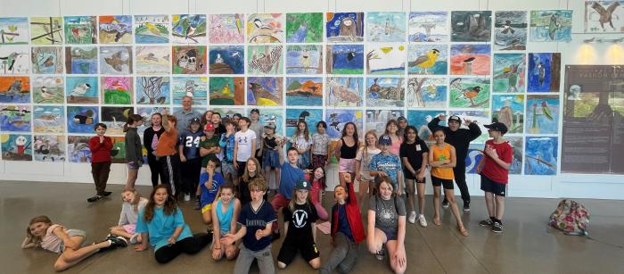 A group of students and one male adult stand in front of a gallery of artwork.