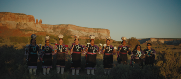 Line of women in ceremonial garb standing with pottery on their heads in front of mountains.