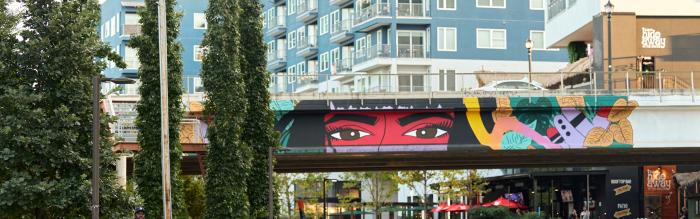 People walk along a paved path with trees to the left and a garden to the right. Behind them, a mural is painted on an overpass, showcasing bright colors and what appears to be a woman's eyes looking out over everything. Behind it is a cluster of buildings including apartments and restaurants.