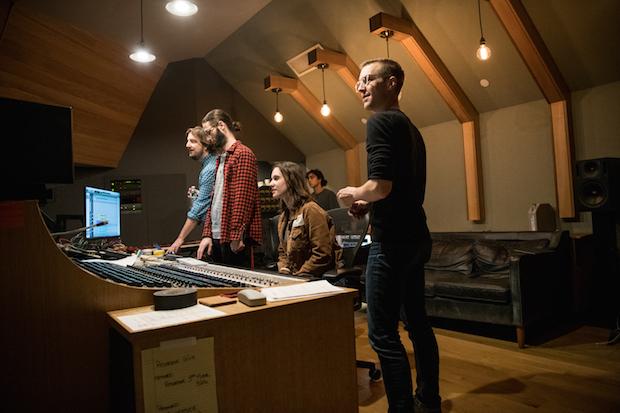 A young women sits in a desk chair in front of a large soundboard in a recording studio, looking at a computer monitor. Standing around her are three men, adjusting the controls. In the background, beside a couch, stands a young man with a ponytail lookin