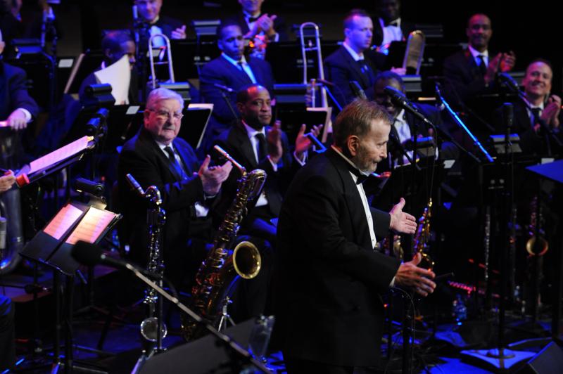 A man looks out at the audience from the stage with an orchestra behind him.