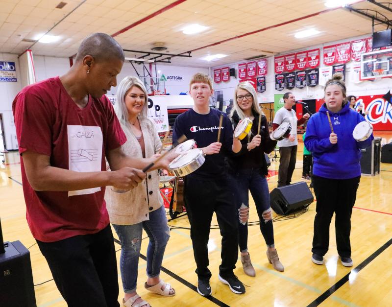 A man shows a group of teenagers in a high school gym how to play a Brazilian instrument