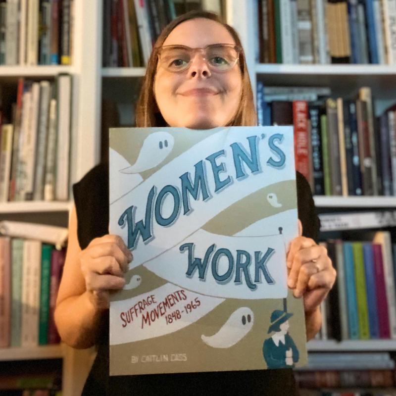 Caitlin Cass A woman with glasses holds up a book that says Women's Work