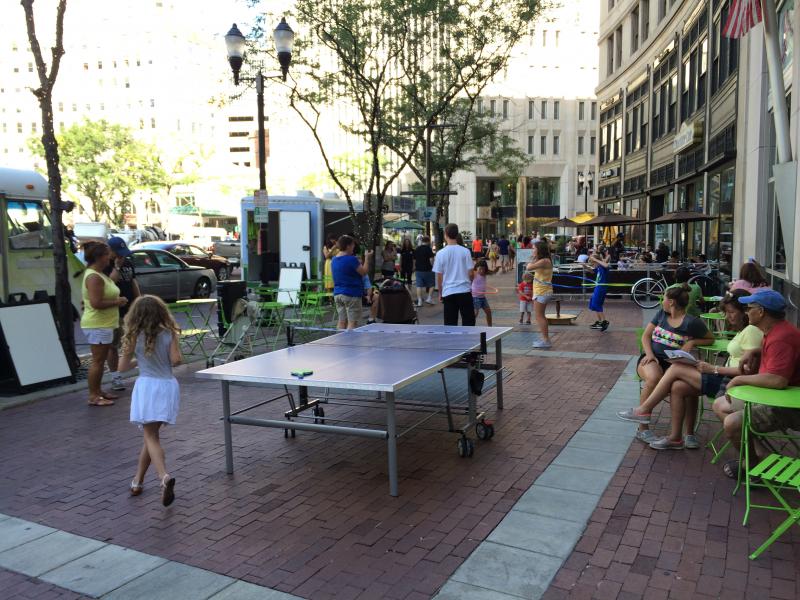 People on the sidewalk of a city doing hula hoop near a ping-pong table.