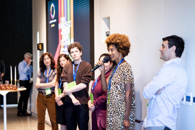 Six people standing up engaged in conversation, as one Black woman holds a mic. Behind the group, is a multi-colored sign that says “SAN FRANCISCO JEWISH FILM FESTIVAL 43.”