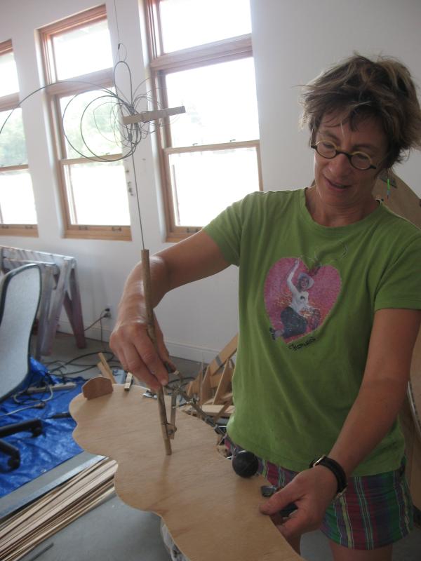 Woman with short brown hair and glasses, wearing a green tee-shirt, working on a sculpture. 
