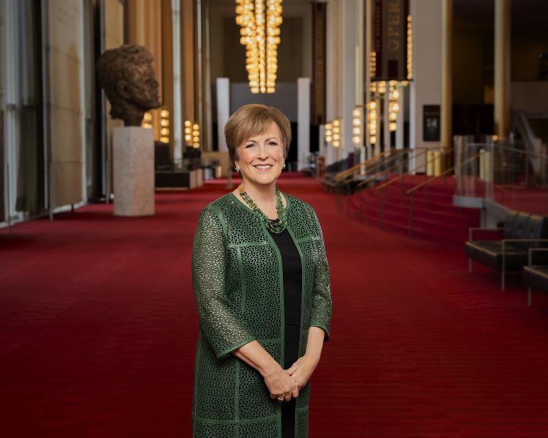 photo of Deborah F. Rutter, a middle-aged white woman with short brown hair. She wears a green loosely woven long jacket over a black dress and stands in the main hall of the Kennedy Center