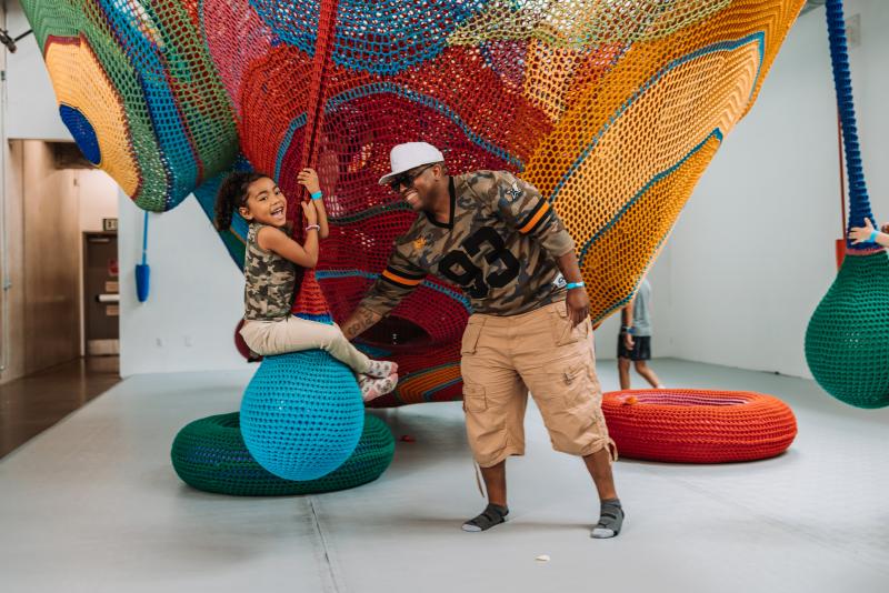 A father plays with his laughing daughter who is swinging on a giant hammock, which is an interactive exhibit at a children's museum
