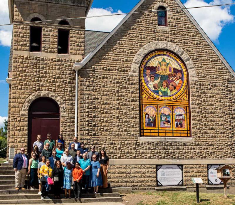 A group of people post on the steps of a stone church. A large window is covered with a mural made of mosaic pieces picturing moments in the history of the church.