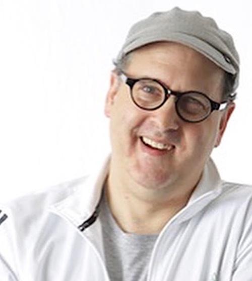 Anthony Ptak is a Caucasian man with clean-shaven, cropped short fine sandy brown hair, high forehead, in his late forties wearing a gray short brimmed cap, glasses with black rims, and a white open zip athletic long sleeve jacket with grey stripes, and a grey cotton heather grey t-shirt underneath. Anthony is smiling asymmetrically and his left eye is slightly rolled back.