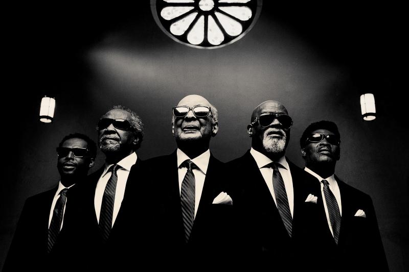 Blind Boys of Alabama, a group of five African-American men standing in a “V” formation. They are wearing black suits ties that display a striped diagonal pattern and black rectangular sunglasses. There is a white pocket square in the front upper left pocket of the jackets of the three men from the middle to the right. There is a lamp on each side of the group and flower-shaped stained glass above them. It is a black and white photograph.